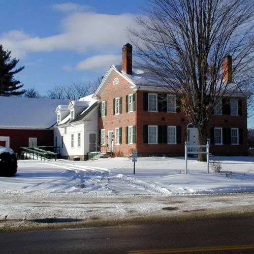 A historic brick building with green shutters, surrounded by snow and trees, on a clear winter day.