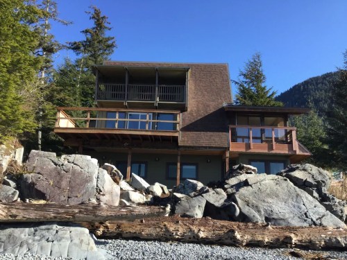 A modern house with multiple balconies, surrounded by large rocks and trees, set against a clear blue sky.