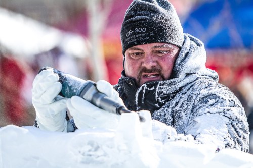 A focused artist carves ice with a tool, wearing a winter coat and gloves, surrounded by a festive outdoor setting.