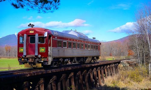 A vintage red train on a wooden trestle, surrounded by autumn foliage and mountains under a blue sky.
