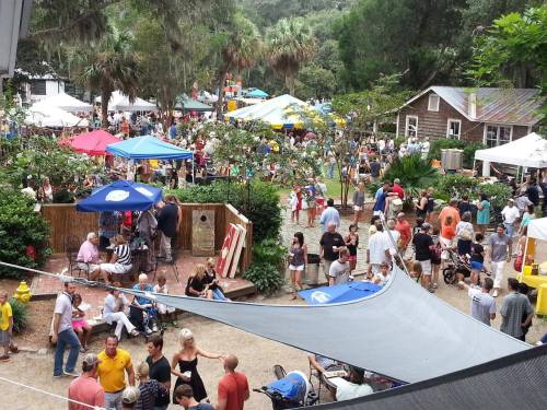 A bustling outdoor festival scene with colorful tents, crowds of people, and greenery in a lively atmosphere.