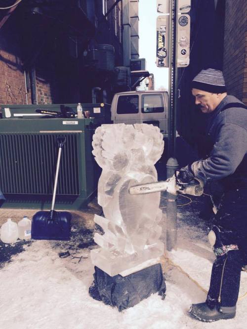 A man carves an ice sculpture in an alley, using a chainsaw, with snow and tools scattered around.
