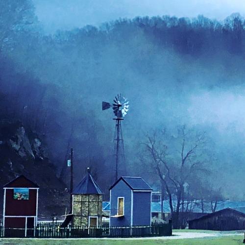 A foggy landscape featuring a windmill and small buildings, surrounded by trees and hills.