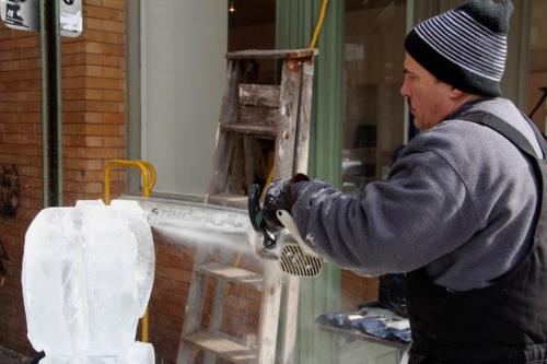 A man carves ice with a chainsaw, focused on his work, with a ladder and building in the background.