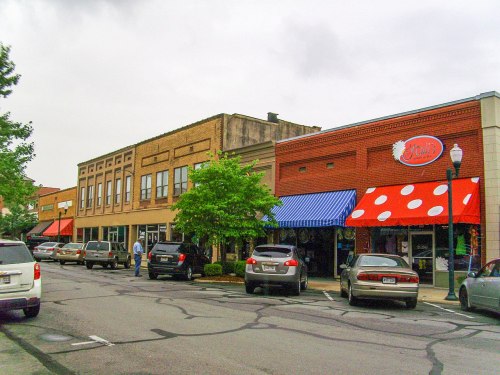 A street view of shops with colorful awnings, parked cars, and trees lining the sidewalk on a cloudy day.