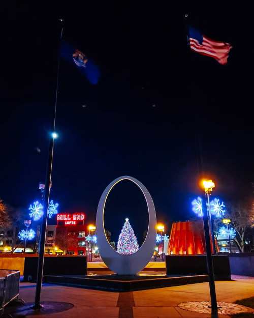 A nighttime scene featuring a large oval sculpture, Christmas tree, and festive lights, with flags in the foreground.