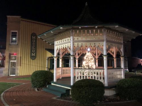 A lit gazebo with a Christmas tree at night, next to a museum building.
