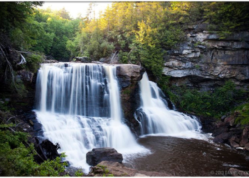 A serene waterfall cascades over rocky cliffs, surrounded by lush green trees and a calm river below.