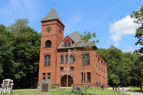 Historic red brick building with a clock tower, surrounded by trees and blue sky.