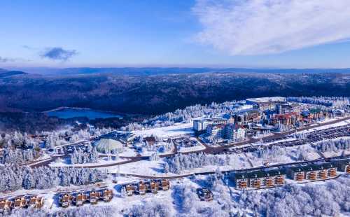 Aerial view of a snowy landscape with a resort, trees, and a lake in the background under a clear blue sky.