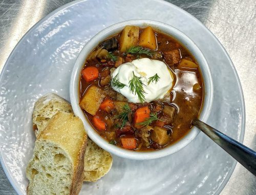 A bowl of hearty vegetable soup topped with sour cream and dill, served with slices of crusty bread on the side.