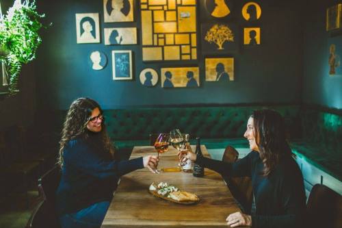 Two women clink glasses of wine at a wooden table, with a stylish wall of silhouettes in the background.