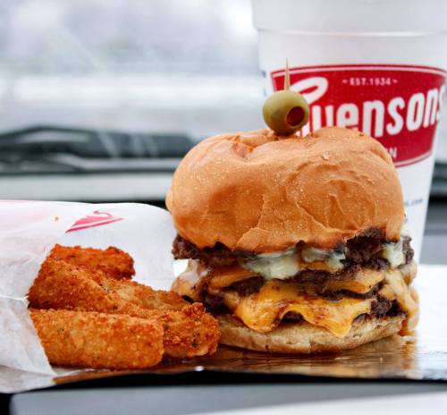 A juicy cheeseburger with two patties and an olive on top, accompanied by crispy golden fries and a drink.