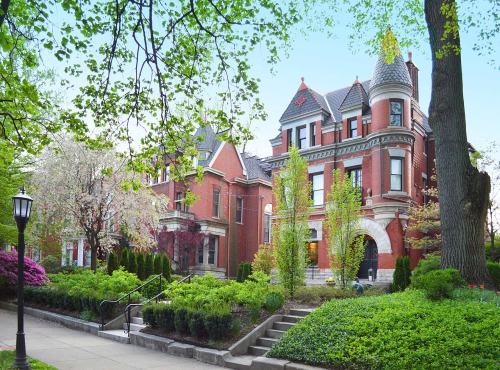 A historic red brick mansion with a turret, surrounded by lush greenery and blooming trees in a serene neighborhood.
