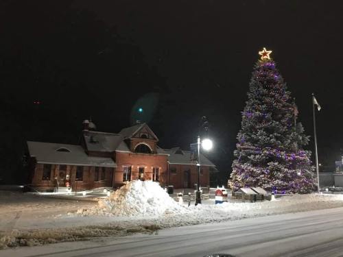 A snowy night scene featuring a large decorated Christmas tree next to a brick building, with snow piled nearby.
