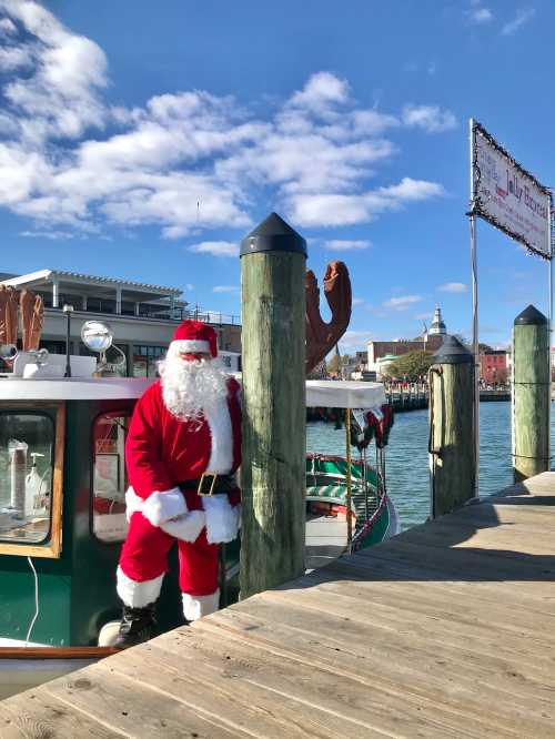 Santa Claus stands by a boat at a dock, with festive decorations and a clear blue sky in the background.