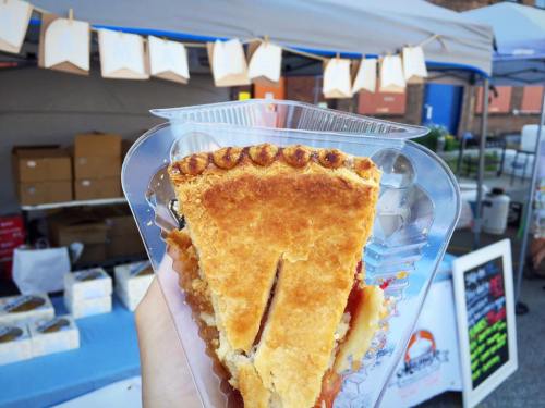 A hand holding a slice of pie in a clear container, with a market stall and decorations in the background.