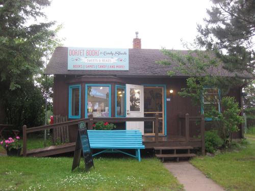 A small brown building with a blue bench, featuring a sign for "Dorset Book & Candy Shoppe" and surrounded by greenery.