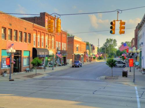 A quiet small-town street lined with shops, American flags, and traffic lights under a clear blue sky.