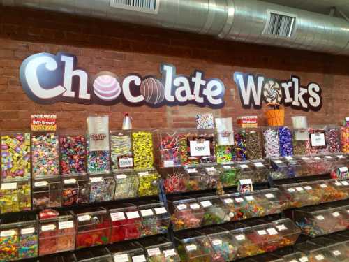 A colorful candy display at Chocolate Works, featuring various sweets in clear bins against a brick wall.