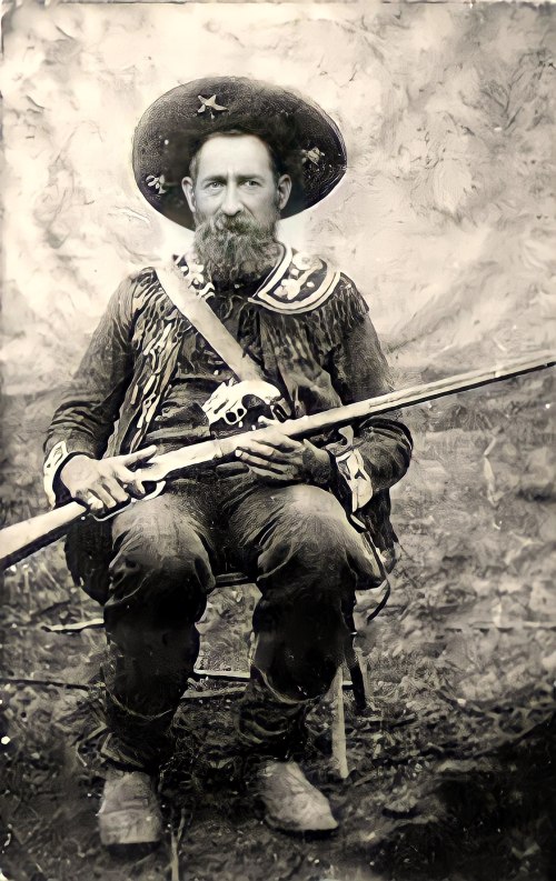 A seated man in a sombrero and fringed clothing holds a rifle, posing against a blurred background.