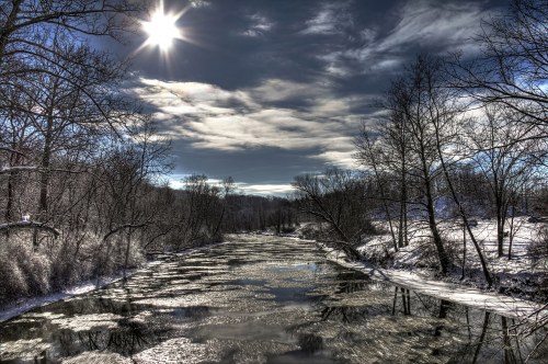 A serene winter landscape featuring a river reflecting the sun, surrounded by snow-covered trees and a cloudy sky.