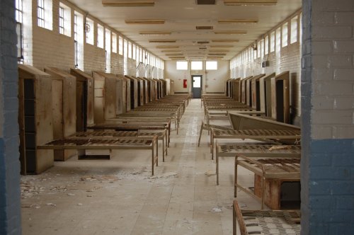 Abandoned prison cell block with empty metal beds and peeling walls, illuminated by natural light from large windows.