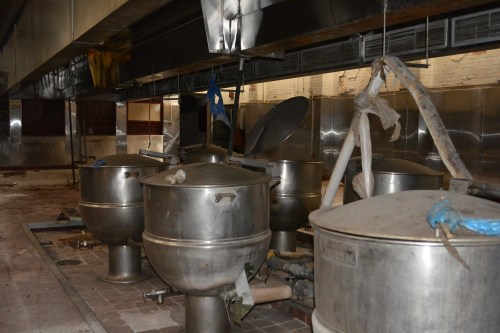 Abandoned kitchen with large metal cooking pots and debris scattered on the floor.