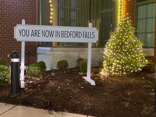 Sign reading "You are now in Bedford Falls" next to a small, lit Christmas tree outside a building.