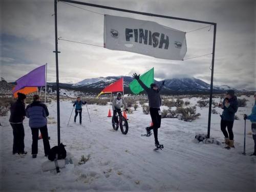 A person celebrates at the finish line of a snowy race, surrounded by flags and spectators in a mountainous landscape.