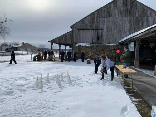 People gather outside a barn in the snow, participating in activities and enjoying a winter event.