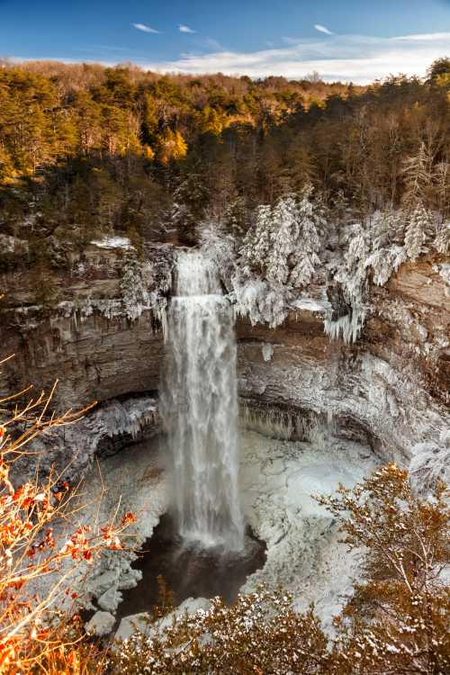 A frozen waterfall surrounded by snow-covered trees and rocky cliffs, capturing a serene winter landscape.