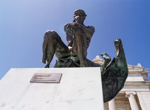 A bronze statue of a seated figure, deep in thought, against a clear blue sky and a classical building backdrop.