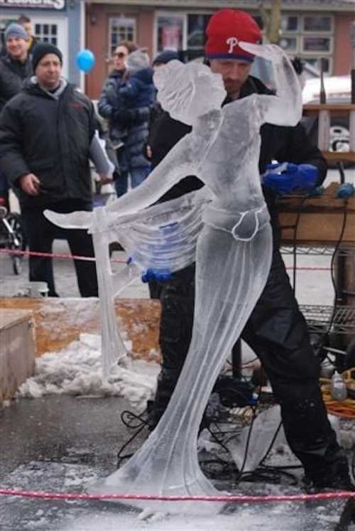 A sculptor carves an ice statue of a woman in a flowing dress at an outdoor event, with onlookers in the background.