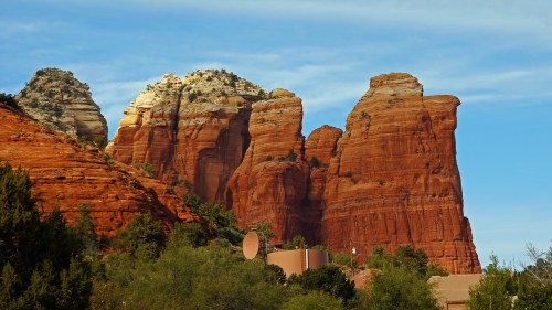 Red rock formations rise against a blue sky, with greenery in the foreground and a building partially visible at the base.