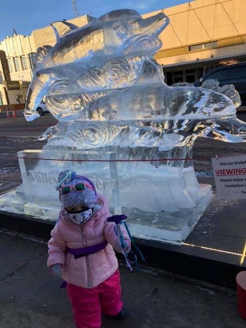 A child in winter clothing stands next to a large ice sculpture, holding a toy, with a city backdrop.