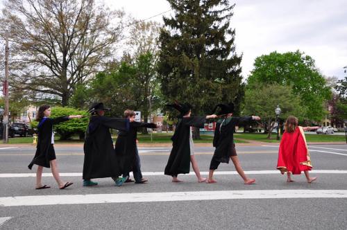 A group of children in costumes walking in a line across a street, with trees and buildings in the background.
