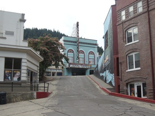 Historic theater building on a sloped street, surrounded by trees and brick buildings, with a mural on the side.