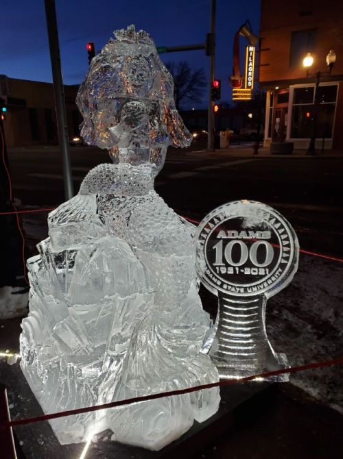 Ice sculpture of a historical figure with a commemorative plaque, illuminated at night in a city setting.