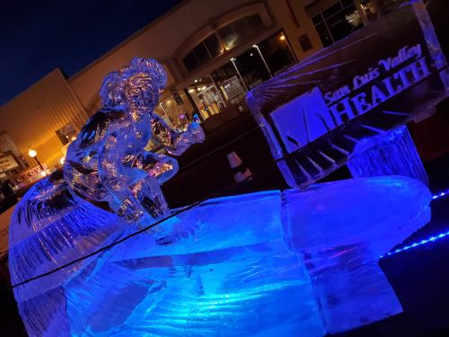 An illuminated ice sculpture of a figure in motion, with a sign for San Luis Valley Health in the background.