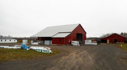 A red barn with a metal roof, surrounded by construction materials, on a cloudy day in a rural area.