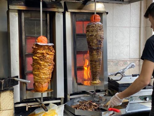 Two vertical spits of meat cooking over flames, with a chef preparing sliced meat on a grill below.