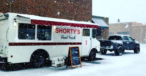 A food truck named "Shorty's" parked in a snowy street, with a truck beside it and snowflakes falling.