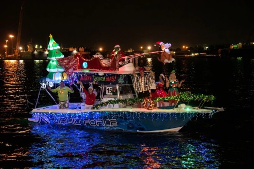 A festive boat decorated with Christmas lights, a tree, and holiday figures, sailing on a dark waterway at night.