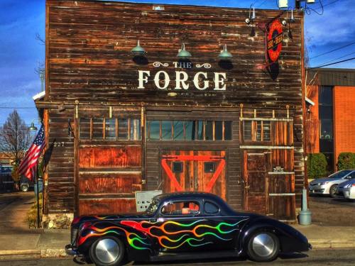A vintage car with flame decals parked in front of a rustic building labeled "The Forge." American flag visible.