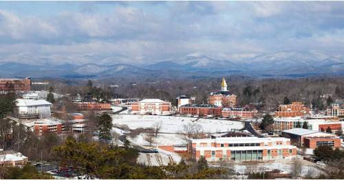 A snowy campus landscape with buildings and mountains in the background under a cloudy sky.