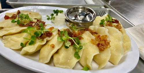 A plate of dumplings topped with crispy bacon and microgreens, served with a small dipping sauce.