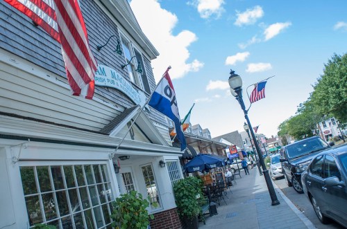 A street view of a pub with flags, outdoor seating, and shops under a blue sky with scattered clouds.