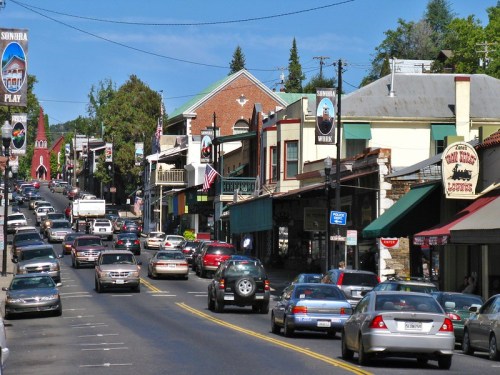 A busy street in a small town with shops, cars, and trees under a clear blue sky.