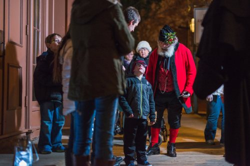 A child looks up at a man dressed as Santa Claus, surrounded by a small crowd in a festive outdoor setting.